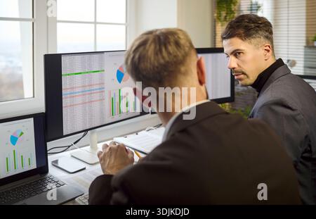 Zwei Analysten prüfen Finanzdaten und Diagramme auf mehreren Büromonitoren Stockfoto