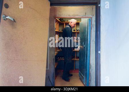 Ein Mann in schwarzen Overalls öffnet eine blaue Tür zu einem Weinlagerbereich, enthüllt Regale mit Flaschen, die eine Atmosphäre der Vorfreude schaffen Stockfoto