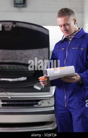 Erwachsener männlicher Mechaniker im Overall, der die Zwischenablage überprüft, orangefarbener Stift in der Garage im silbernen Auto, Kopierraum. Techniker, Motor, Inspektion, Checkliste, Dokumentation Stockfoto