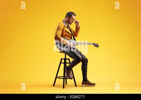 Stilvoller männlicher Musiker, der elektrische Gitarre spielt und auf einem Hocker vor einem leuchtend gelben Hintergrund sitzt Stockfoto