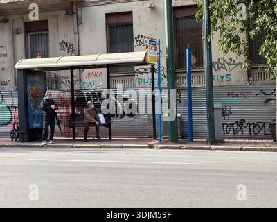 Bushaltestelle in Athen, Griechenland (Αθήνα). Urbane Szene mit Passagieren, einer trägt eine Gesichtsmaske, vor einer Hintergrundwand, die mit lebhaften Graffiti A bedeckt ist Stockfoto