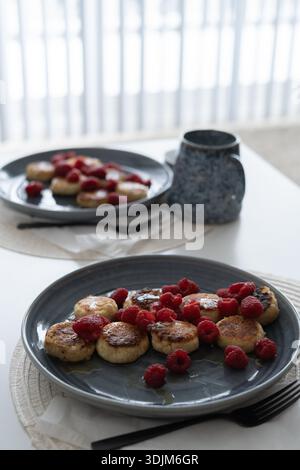 Eine gemütliche Frühstücksszene mit zwei Tellern traditioneller slawischer Syrniki mit Hüttenkäse, mit Honig getränkt und mit frischen Himbeeren belegt Stockfoto