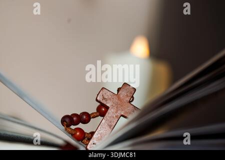 Ein Rosenkranz liegt auf den Seiten einer offenen Bibel. Eine flackernde Kerze im Hintergrund sorgt für eine warme Atmosphäre im Raum. Leuchte leuchtet auf Stockfoto