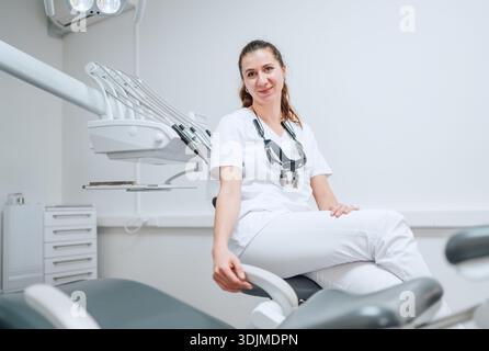 Portraait einer aufrichtig lächelnden jungen Zahnarztfrau gekleidet weißen medizinischen Peelings Uniform sitzend in der modernen Zahnklinik neben dem Stomatologie-Stuhl. Hea Stockfoto