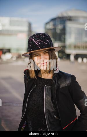 Ein stilvolles Kopfbild einer jungen, modischen Frau mit Hut, T-Shirt und Bomberjacke aus schwarzem Leder in urbanem Ambiente. Stockfoto