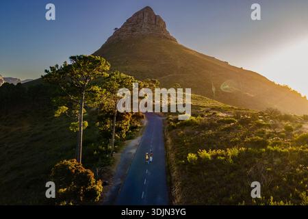 Zwei Wanderer spazieren entlang einer gewundenen Straße, die zum Lions Head führt, umgeben von üppigem Grün. Der goldene Sonnenuntergang strahlt ein warmes Leuchten über dieser legendären Landma aus Stockfoto