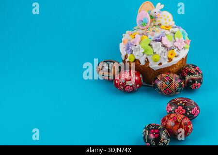 Ein runder Kuchen sitzt auf einer hellblauen Oberfläche. Der Kuchen wird mit Glasur, Süßigkeiten und farbenfrohen Dekorationen gekrönt. Um den Kuchen herum sind Eier in verschiedenen Stücken dekoriert Stockfoto