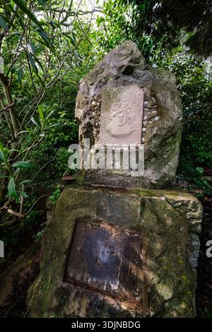 Steindenkmal mit einem geschnitzten Relief eines Gesichts in einem großen Felsen, umgeben von üppigen grünen Bäumen und Büschen in einer waldähnlichen Umgebung. Stockfoto