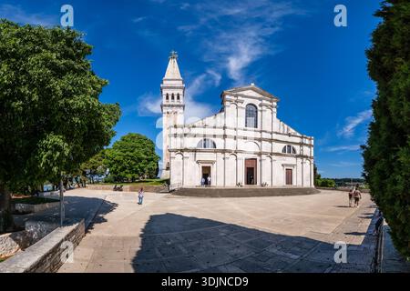 Vorderansicht der Kirche St. Euphemia in Rovinj unter blauem Himmel, mit Besuchern auf dem umliegenden Steinplateau und Bäumen an den Seiten. Stockfoto