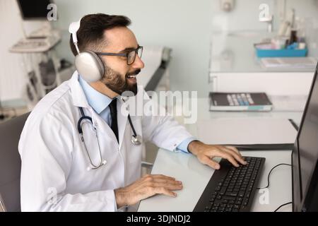 Lächelnder junger männlicher Arzt in digitalen Kopfhörern, die am Computer arbeiten Stockfoto