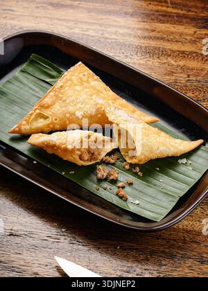 Eine Nahaufnahme von drei goldbraunen Samosas, eines aufgebrochen, um eine herzhafte Füllung zu enthüllen, präsentiert auf einem frischen grünen Bananenblatt auf einem Holztisch. Stockfoto