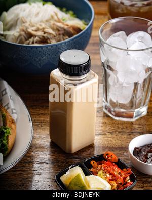 Ein erfrischendes Eiskaffee in einer modernen quadratischen Flasche steht auf einem Holztisch. Die Szene enthält auch eine Schüssel vietnamesischer Pho-Nudeln, ein g Stockfoto