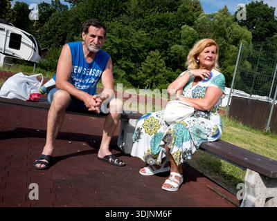 Ein älterer Mann und eine Frau sitzen an einem sonnigen Tag auf einer Parkbank. Der Mann trägt ein blaues Tanktop und die Frau trägt ein Blumenkleid mit einer Geldbörse Stockfoto