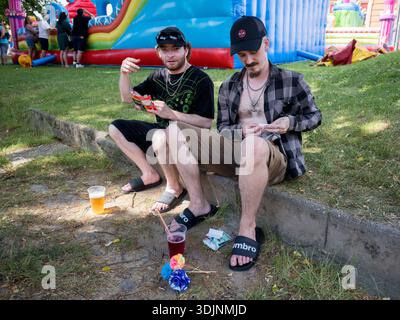 Zwei junge Männer sitzen auf einem Bordstein auf einem Outdoor-Festival, Rollen Zigaretten und trinken. Ein buntes aufblasbares Hüpfschloss im Hintergrund Stockfoto