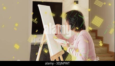 Malerin mit rosafarbenem Oberteil und Schürze zu Hause mit Staffelei und Palette, Pinsel, Grafiken Stockfoto