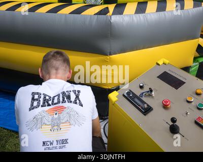 Ein Mann in einem weißen Bronx-T-Shirt bedient auf einem Jahrmarkt ein Bedienfeld für eine aufblasbare Vergnügungsattraktion mit gelben und schwarzen Gefahrenstreifen. Stockfoto
