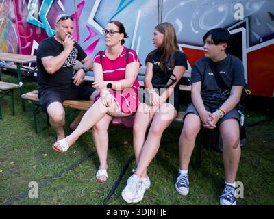 Eine vierköpfige Familie sitzt auf einer Holzbank auf einem Freiluftmarkt, ein Mann in einem schwarzen T-Shirt raucht und eine Frau in einem roten Kleid blickt nach vorne Stockfoto