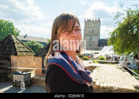 Eine junge Frau lächelt in einem historischen Innenhof, mit alten Backsteinbauten und dem imposanten Steinturm von Lubart's Castle, der unter hellem Himmel zu sehen ist. Stockfoto