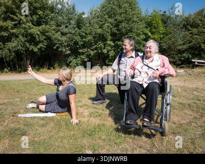 Eine Frau aus den 50er Jahren, die auf dem Gras sitzt, ein Selfie mit einer älteren Frau im Rollstuhl macht und ein Mann aus den 60er Jahren, der neben ihr in einem Park kniet. Stockfoto