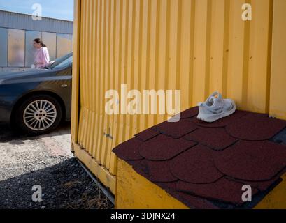 Ein einzelner weißer Sneaker auf dem dunkelroten Dach eines kleinen gelben Holzbaus, mit einer Frau aus den 30er Jahren und einem grauen Auto im unscharfen Hintergrund. Stockfoto