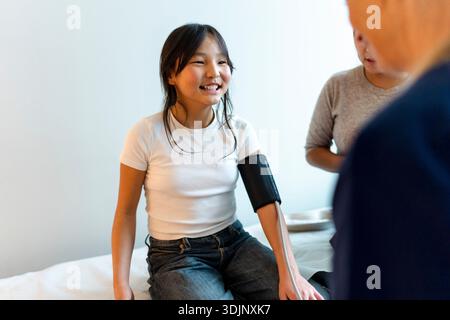 Lächelndes Mädchen mit Blutdruckgürtel, das während des Krankenhausbesuchs auf dem Untersuchungstisch sitzt Stockfoto