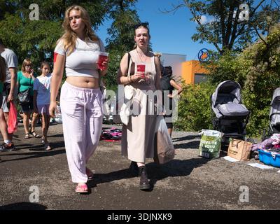 Zwei junge Frauen, die durch einen Flohmarkt laufen und rote Plastikbecher halten. Kinderwagen und verschiedene Waren werden auf dem Gelände in der Nähe ausgestellt. Stockfoto