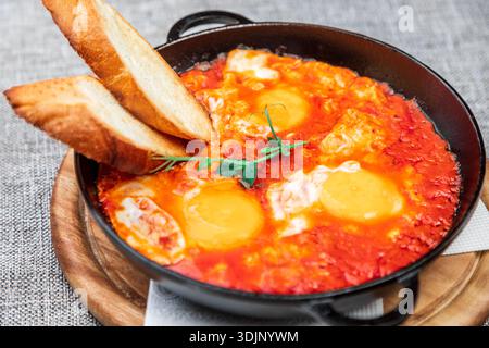 Shakshuka (Eier in einer scharfen Tomatensauce pochiert) mit knusprigen Toasts. Es wird in einer schwarzen gusseisernen Pfanne auf einem Holztablett serviert. Nahaufnahme Stockfoto