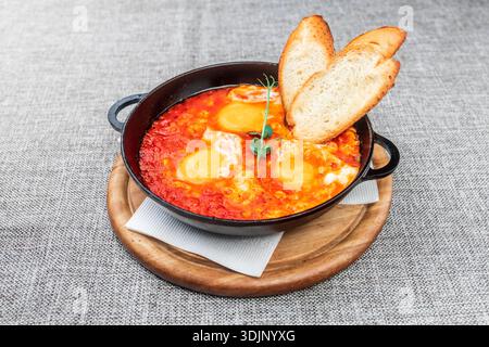 Shakshuka (Eier in einer scharfen Tomatensauce pochiert) mit knusprigen Toasts. Es wird in einer schwarzen gusseisernen Pfanne auf einem Holztablett serviert. Eine graue Tischdecke wie eine Ba Stockfoto