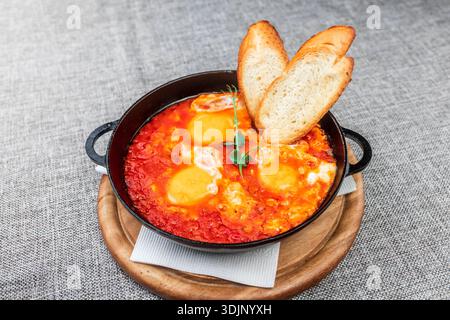 Shakshuka (Eier in einer scharfen Tomatensauce pochiert) mit knusprigen Toasts. Es wird in einer schwarzen gusseisernen Pfanne auf einem Holztablett serviert. Eine graue Tischdecke wie eine Ba Stockfoto