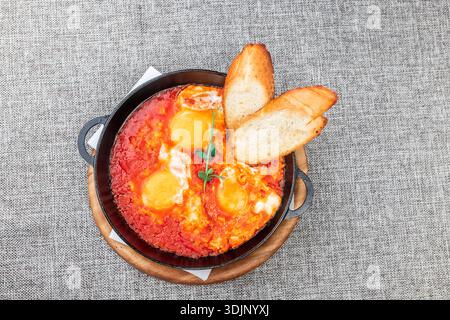 Shakshuka (Eier in einer scharfen Tomatensauce pochiert) mit knusprigen Toasts. Es wird in einer schwarzen gusseisernen Pfanne auf einem Holztablett serviert. Eine graue Tischdecke wie eine Ba Stockfoto