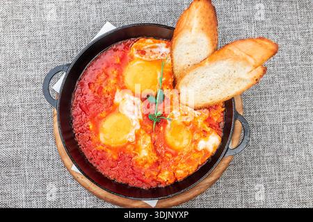 Shakshuka (Eier in einer scharfen Tomatensauce pochiert) mit knusprigen Toasts. Es wird in einer schwarzen gusseisernen Pfanne auf einem Holztablett serviert. Eine graue Tischdecke wie eine Ba Stockfoto