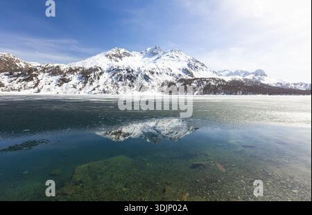 Aus der Vogelperspektive auf den majestätischen, schneebedeckten Berg, der sich im türkisfarbenen Wasser des Sees Sils-Maria, Schweiz, spiegelt. Stockfoto