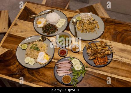 Blick von oben auf ein appetitliches asiatisches Essen auf rustikalem Holztisch. Diese Mahlzeit umfasst gebratenen Reis, gegrillte Satay-Spieße, Fleisch und verschiedene scharfe Dippi Stockfoto