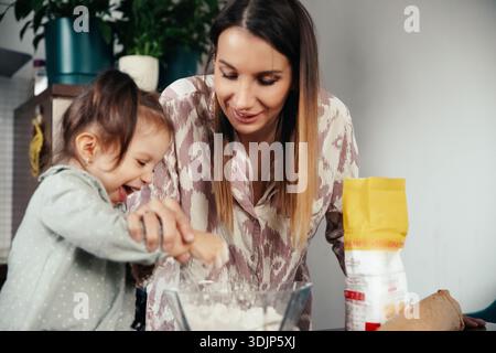 Mutter und Tochter kochen gerne in ihrer Küche. Die Tochter lacht, während sie Mehl in einer Schüssel rührt, während die Mutter glücklich zusieht. Sie teilen einen Stockfoto