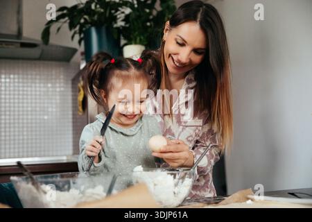 Eine Frau und ein Mädchen sind in einer Küche. Sie lächeln und arbeiten zusammen. Das Mädchen hält ein Ei, während die Frau ihr bei der Zubereitung des Essens hilft Stockfoto