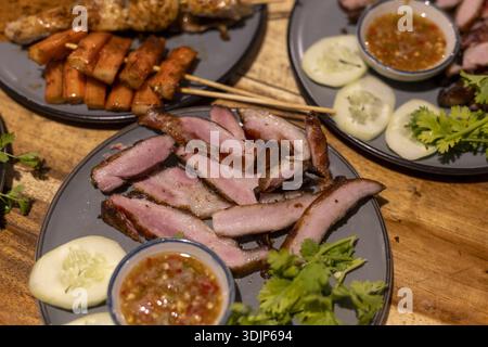 Köstlicher thailändischer gegrillter Schweinehals mit würziger Dippsoße. Herzhafte asiatische Küche, beliebtes Street Food Gericht serviert auf dunklem Teller mit frischer Gurke und Stockfoto