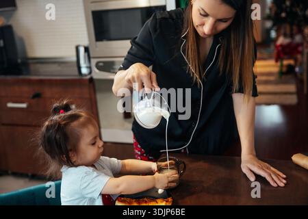Eine Mutter gießt Milch in ihren Kinderbecher am Küchentisch. Das Kind beobachtet genau, begierig auf den Drink. Die Umgebung zeigt einen warmen Familienmorgen Stockfoto