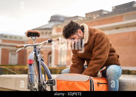 Junger Mann, der als Fahrradkurier arbeitet und die Thermotasche öffnet, um einen Lieferauftrag in einer städtischen Umgebung zu überprüfen Stockfoto