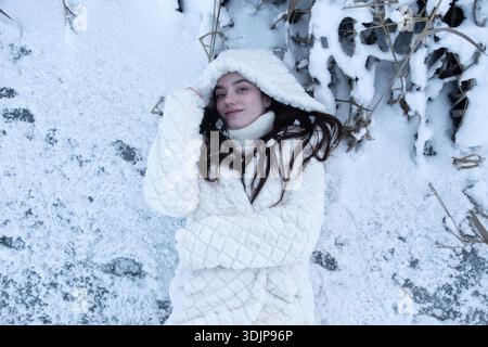 Junge Frau, die im Neuschnee liegt und einen weißen Wintermantel und eine Kapuze trägt, die entspannt und zufrieden von oben aussieht und Wärme, Intimität, Kontrast und Qu ausdrückt Stockfoto