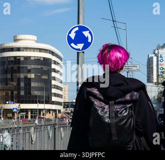 Kiew, Ukraine - 6. Mai 2023 : Person mit hellrosa Haaren und Rucksack, die an einem Kreisverkehr-Schild auf einer sonnigen modernen Stadtstraße in Kiew, Ukraine, vorbeiläuft Stockfoto