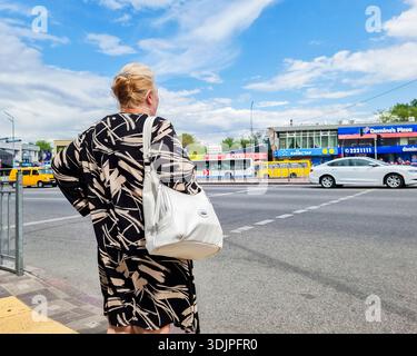 Kiew, Ukraine - 6. Mai 2023 : Frau wartet auf einem Kiewer Bürgersteig, zurück zur Kamera, beobachtet den Verkehr und das tägliche Leben in der Stadt unter einem Sommerhimmel Stockfoto