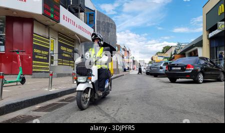 Kiew, Ukraine - 6. Mai 2023 : Lieferfahrer in gelber Weste und Helm auf einem Roller, der durch eine geschäftige Kiewer Straße mit Geschäften und Sommermonaten schlängelt Stockfoto