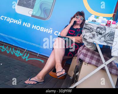 Kiew, Ukraine - 6. Mai 2023 : Frau mittleren Alters sitzt an einem Straßenstand und spricht mit Smartphone neben einem blauen Werbebanner auf einem Kiewer Bürgersteig, CA Stockfoto