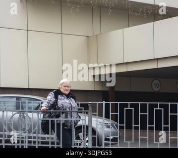 Kiew, Ukraine - 6. Mai 2023 : ältere Frau in Winterjacke steht am Metallzaun auf einer städtischen Kiewer Straße, besinnlicher Ausdruck vor Moder Stockfoto
