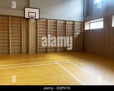 Der Basketballkorb befindet sich an einer Holzwand in einer Schulsporthalle mit Holzböden und Regalen, wobei Sonnenlicht durch den Sieg strömt Stockfoto