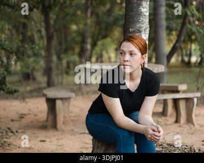 Foto einer jungen Frau, die auf einer Steinbank in einem Park sitzt, sich nach vorne beugt und zur Seite blickt. Angemessenes Bild für Reflexionskonzepte, Stockfoto