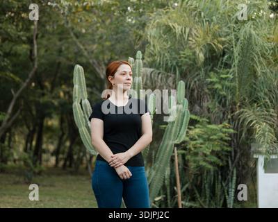 Bild einer jungen Frau, die in einem Park steht, mit Kakteen und Vegetation im Hintergrund, mit entspannter Haltung und Blick zur Seite Stockfoto