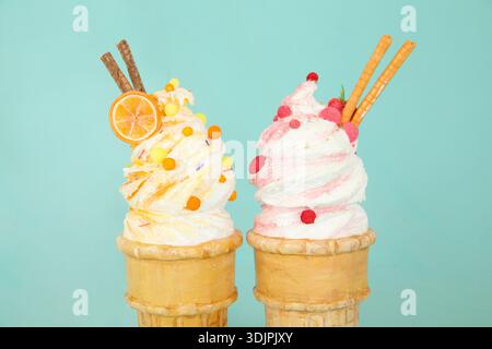 Zwei Eiskegel mit weichen Servierwirbeln, einer mit einer Orangenscheibe und bunten Streuseln, der andere mit Himbeeren und Waffelstäbchen Stockfoto
