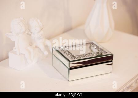 In einem sanft beleuchteten Raum befindet sich neben zwei weißen Cherub-Figuren auf einer Holzoberfläche eine mit Strasssteinen verspiegelte Schmuckschatulle Stockfoto