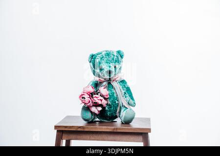 Teddybär mit rosafarbenen Rosen, auf einem Holzhocker platziert, auf einem sauberen weißen Hintergrund, geeignet für verschiedene dekorative Zwecke Stockfoto